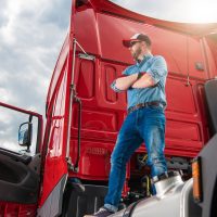 Proud Caucasian Trucker in His 30s and His Brand New Semi Truck Tractor. Truck Driver Staying on Top of the Vehicle Rear End. Heavy Duty Transportation Industry.